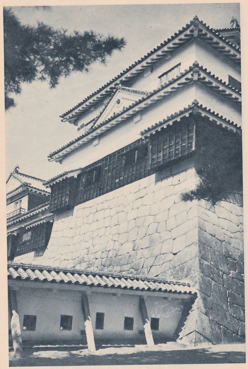 Black-and-white photograph, taken from below, of a white-walled building of stacked tiled roofs with a gable and a dark band of wooden panels that curves out over a large wall of fitted stones. In front at the lower left a long low white wall under a tiled roof, with small square dark windows, is held up by slanting posts. A dark pine branch hangs at the upper left and another shows at the right.