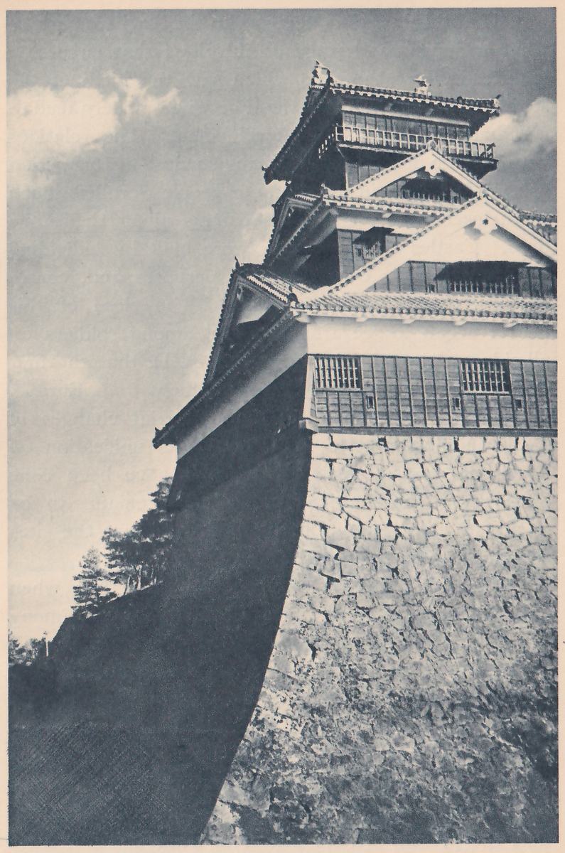Black-and-white photograph of the corner of a tower of stacked tiled roofs, rising from a tall stone wall that curves out at its foot and fills the right and lower part of the picture. The upper stories have pale gabled roofs, a railed balcony and dark boarded walls with small barred windows. The left side of the wall lies in deep shadow, and a few pines show against a cloudy sky at the left.