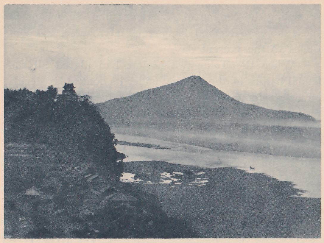 Black-and-white photograph of a wide stretch of water in haze, with a steep tree-covered hill at the left that carries a small building with stacked roofs at its top, and a broad conical mountain across the water at the center. Roofs of a few small buildings show among dark trees at the lower left, and a tiny dark mark lies on the water at the right.