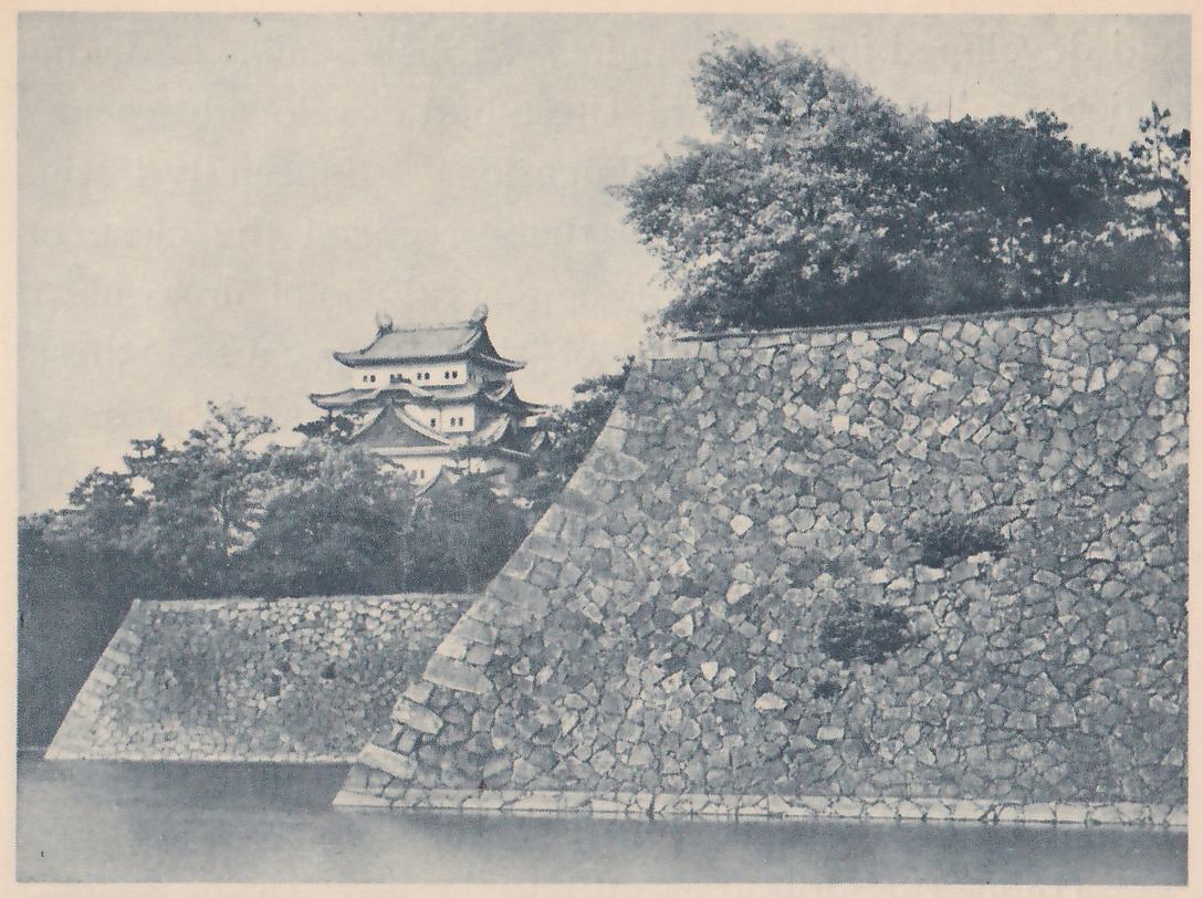 Black-and-white photograph of a sloping stone wall rising straight from still water, with a lower, smaller section of wall at the left in front of trees. A white-walled building with curved dark roofs and a small pointed ornament at each end of its top ridge shows above the trees at the center left. Dense leafy trees crown the large wall at the upper right.