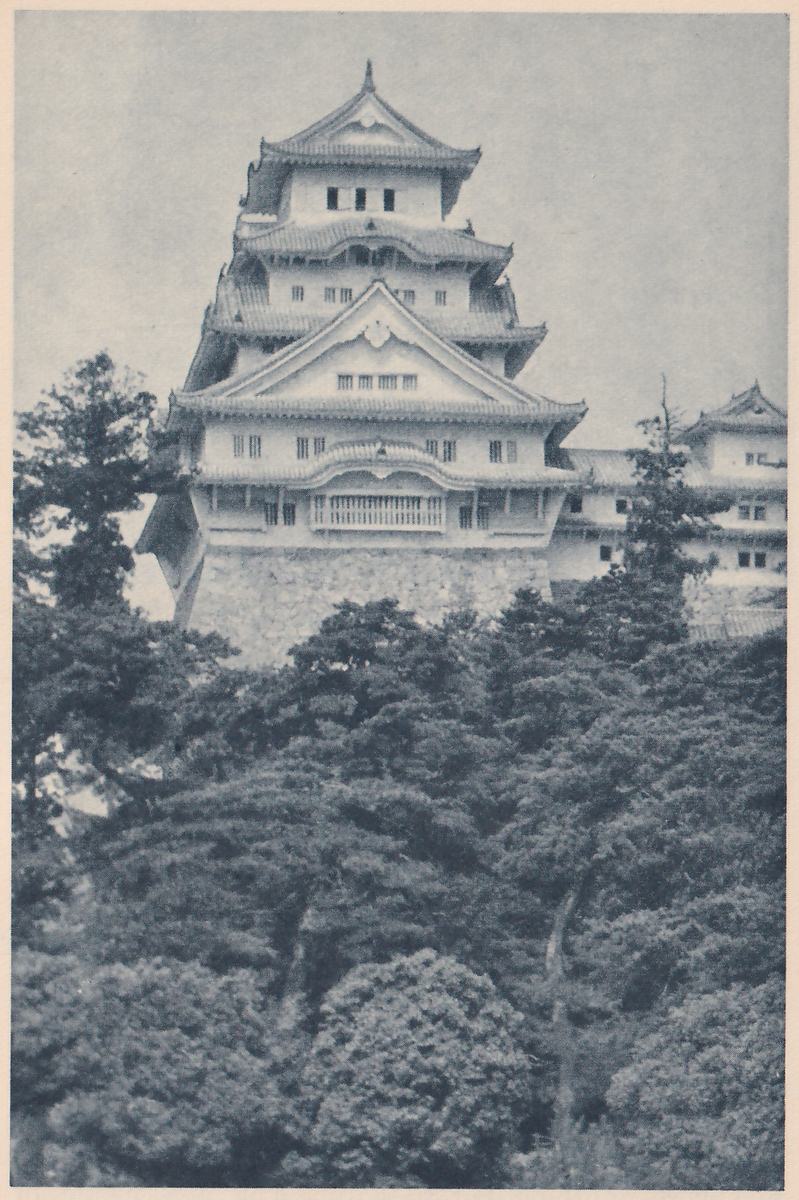 Black-and-white photograph, taken from below, of a tall white building with several stacked tiled roofs, pale gables with small ornaments and rows of small windows, standing on a stone base above dense dark trees that fill the lower half. A lower white building with roofs shows at the right, and a leafy branch crosses the left side.