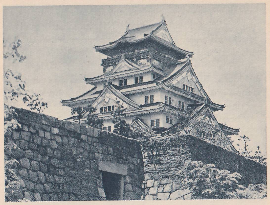 Black-and-white photograph, taken from below, of a white building of stacked tiled roofs with curved eaves, pale gables with fine lattice patterns and rows of small windows. A dark wall of fitted stones, partly covered with leafy growth, runs across the lower half; at the lower left it has a dark doorway-shaped opening under a flat lintel stone. Leaves show at the left edge and the lower right.