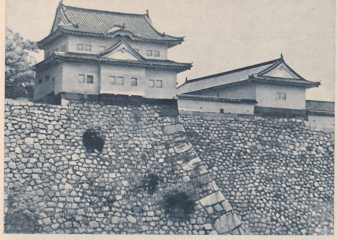 Black-and-white photograph of a steep, sloping wall of fitted stones filling the lower two-thirds of the picture, with a darker round patch near its upper left, tufts of plants in front, and a corner of larger squared stones running diagonally down to the lower right. A white-walled building with two tiled roofs, one above the other, and small square windows stands on top at the upper left, and a long low white-walled building with a gabled end stands at the right.
