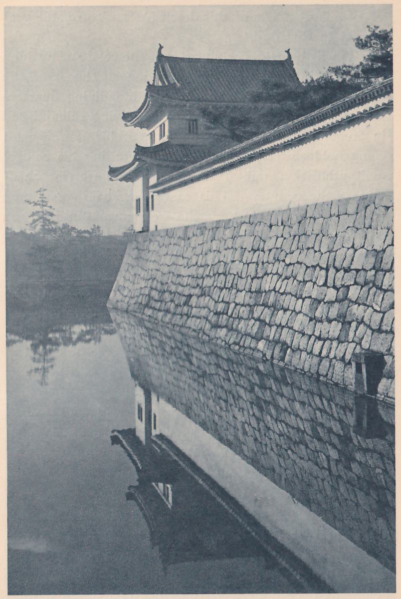 Black-and-white photograph of a white-walled tower with two stepped tiled roofs, the upper one with a pointed ornament at each end of its ridge, beside a long white wall with a tiled top that runs toward the right above a wall of fitted stones. The stone wall and the tower are reflected in still water that fills the lower half of the picture. A pine tree and low trees show at the left, and more trees at the upper right.