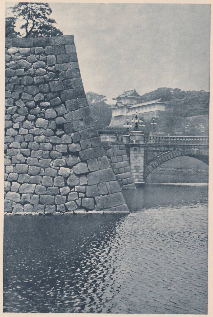 Black-and-white photograph of a large sloping wall of fitted stones at the left, rising from dark rippled water. At the center right a stone bridge with a balustrade and lamp posts crosses the water, with an arch under its right part. Behind the bridge a white-walled building with a tiled upper roof stands on a stone base among trees. A pine shows at the upper left.