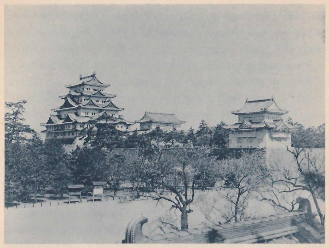 Black-and-white photograph across a wide pale open space with a few small bare-branched trees and a few low boards on posts, toward a line of dark trees. Above the trees stand, from left to right, a tall building of stacked roofs, a lower building with a long tiled roof, and a white-walled building with two roofs at the edge of a wall. The edge of a tiled roof with a curled end appears at the lower right foreground.