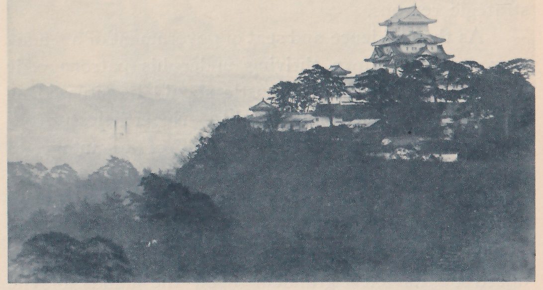 Black-and-white photograph of a wooded hill rising from haze, with a building of stacked roofs at its top right and smaller roofed buildings below it to the left, among pine trees and pale wall segments. Misty hills and two thin upright shapes show far off at the left. Dark treetops fill the lower half.