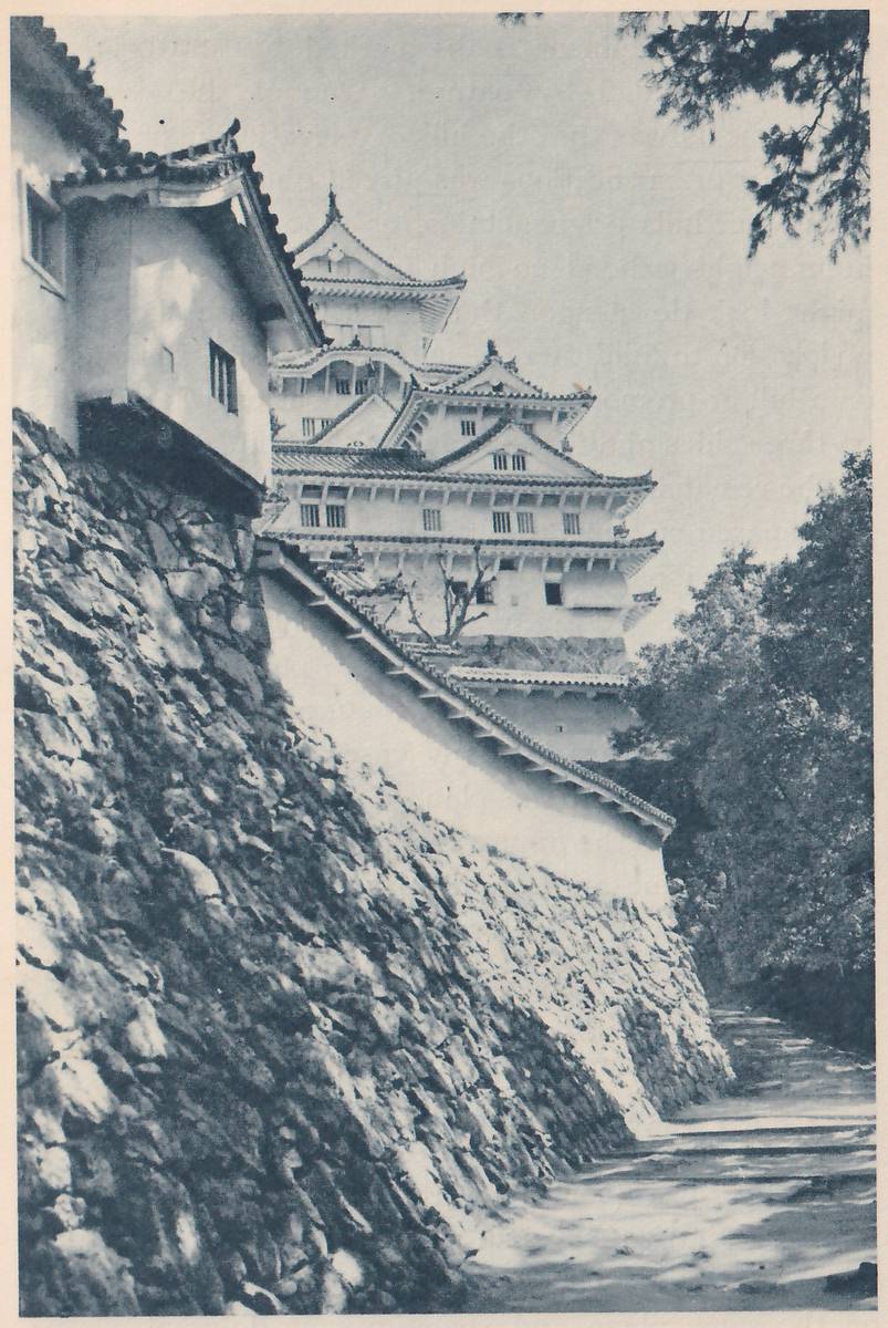 Black-and-white photograph looking up a steep slope of fitted stones at the left, with a white-walled building at its top left corner. A white wall with a tiled top runs along the slope toward the center, and beyond it a white-walled building of stacked tiled roofs with small gables and rows of small windows rises against the sky. Dark leaves hang at the upper right, trees fill the right edge, and a pale sloping path runs along the foot of the slope at the lower right.