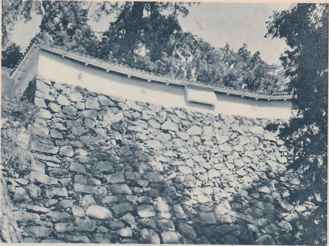Black-and-white photograph, taken from below, of a sloping wall of rough stones, light and shadowed, filling most of the picture, with a long white wall with a tiled top above it. A small square panel stands out from the white wall at the center right. Dark trees rise above the wall and at the right edge.