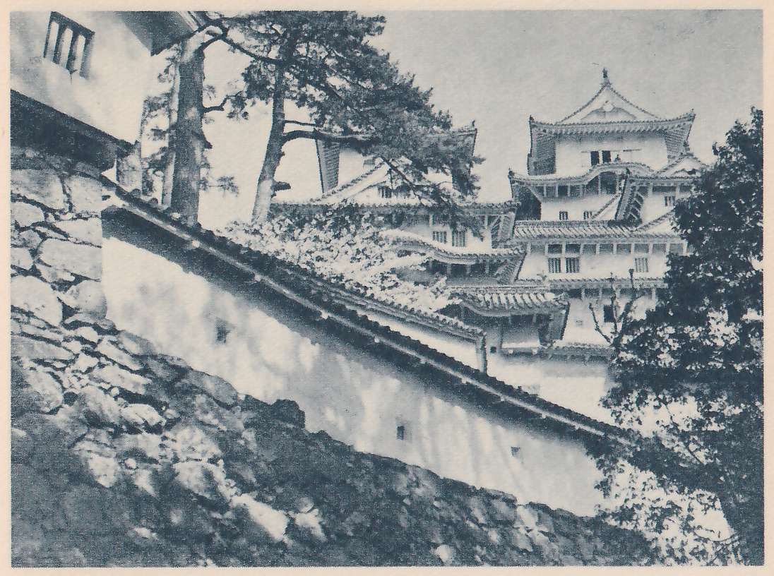 Black-and-white photograph looking up past a sloping stone wall at the lower left. A white wall with a tiled top and a few small dark square openings runs diagonally across the middle toward a white-walled building of stacked tiled roofs with many small windows at the upper right. Pine branches arch over the center and dark leafy trees stand at the right. A white-walled building with a barred window shows at the top left corner.