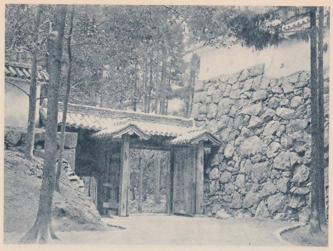 Black-and-white photograph of a wooden gate with a small tiled roof and a wooden door standing in a gap between a bank of earth at the left and a high wall of fitted stones at the right. Tall slender tree trunks and a pale path lie in front. A white wall with a tiled top stands above the stones at the upper right, and a tiled roof edge shows at the left.