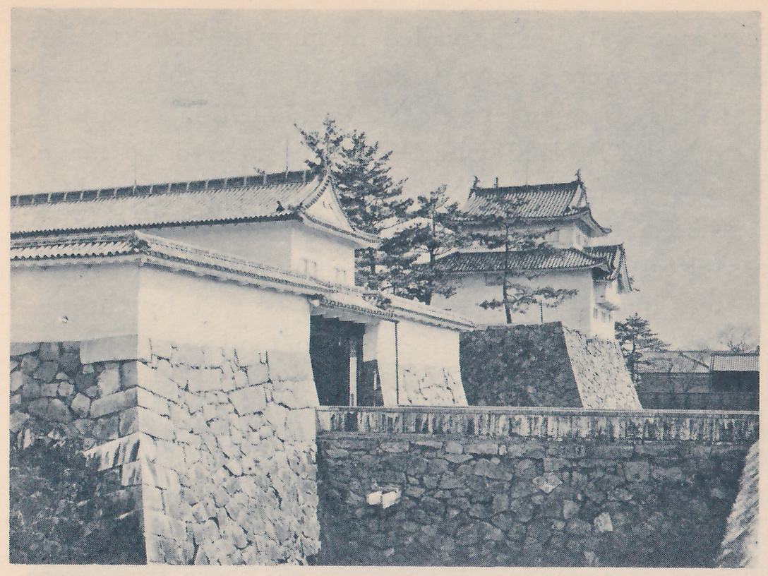 Black-and-white photograph of a white-walled gate building with a long tiled roof, its dark passage opening toward the center, standing on a base of fitted stones. Beside it at the right a white-walled building with two stepped tiled roofs stands on a stone base behind pine trees. A low pale-topped wall runs across the front above a dark stone wall in the lower right.