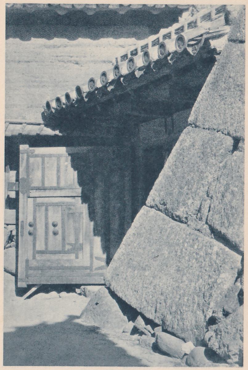 Black-and-white photograph of a gate passage in deep shadow under a tiled eave whose row of round tile ends runs diagonally across the top. A wooden door with framed panels and round studs stands at the left. At the right a wall of very large rough stones slants steeply, with loose stones at its foot and a pale sunlit path at the lower left.