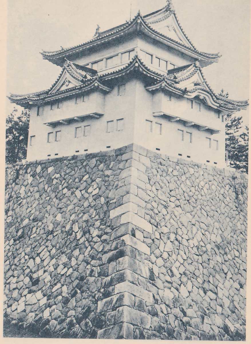 Black-and-white photograph of a white-walled building with stepped tiled roofs, the upper ones with gables and curved eaves, rows of small square windows and shelf-like wooden projections from the wall. It stands on a tall wall of fitted stones whose squared corner stones climb the center in a ridge. Pine trees show at the left and right edges.