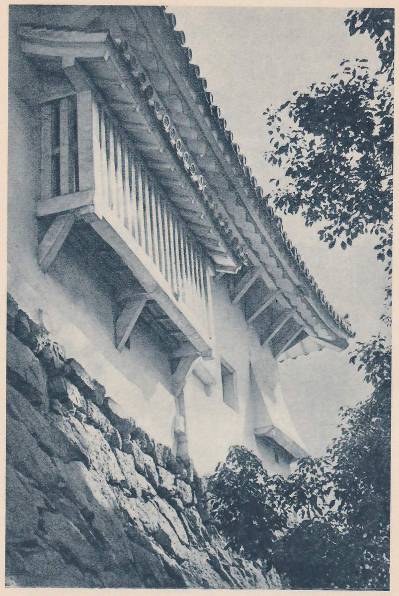 Black-and-white photograph, looking up, of a white-walled box-shaped part that juts out from the wall on slanting wooden brackets, with a row of tall vertical slats across its front and a small window in its left side, under a tiled roof with round tile ends. A small window and a small hood-like roof show lower down on the white wall. A rough stone wall fills the lower left and leaves fill the right.