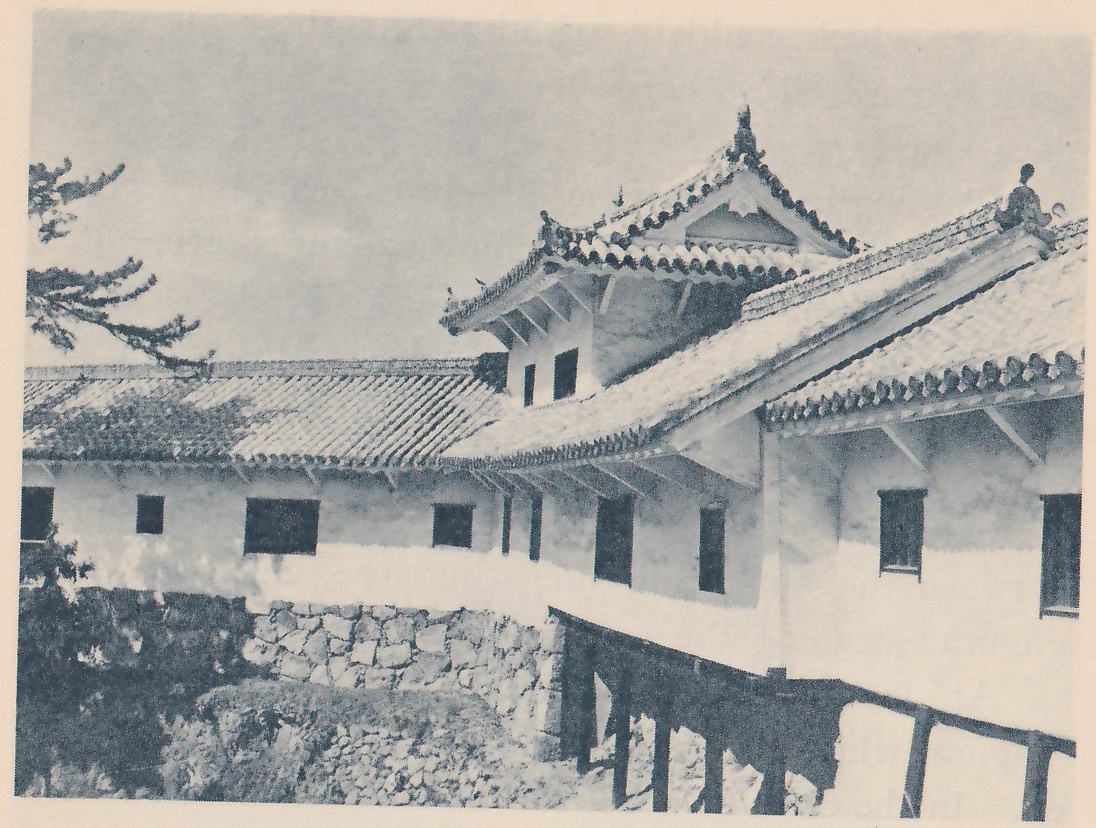 Black-and-white photograph of long white-walled buildings with tiled roofs around a low pale stone wall and a bush at the lower left. A small gabled roof with a pointed ornament rises at the center, and the long roof at the right carries an ornament at its upper end. Dark rectangular windows show along the white walls, and wooden posts cross the lower right. A pine branch shows at the left.