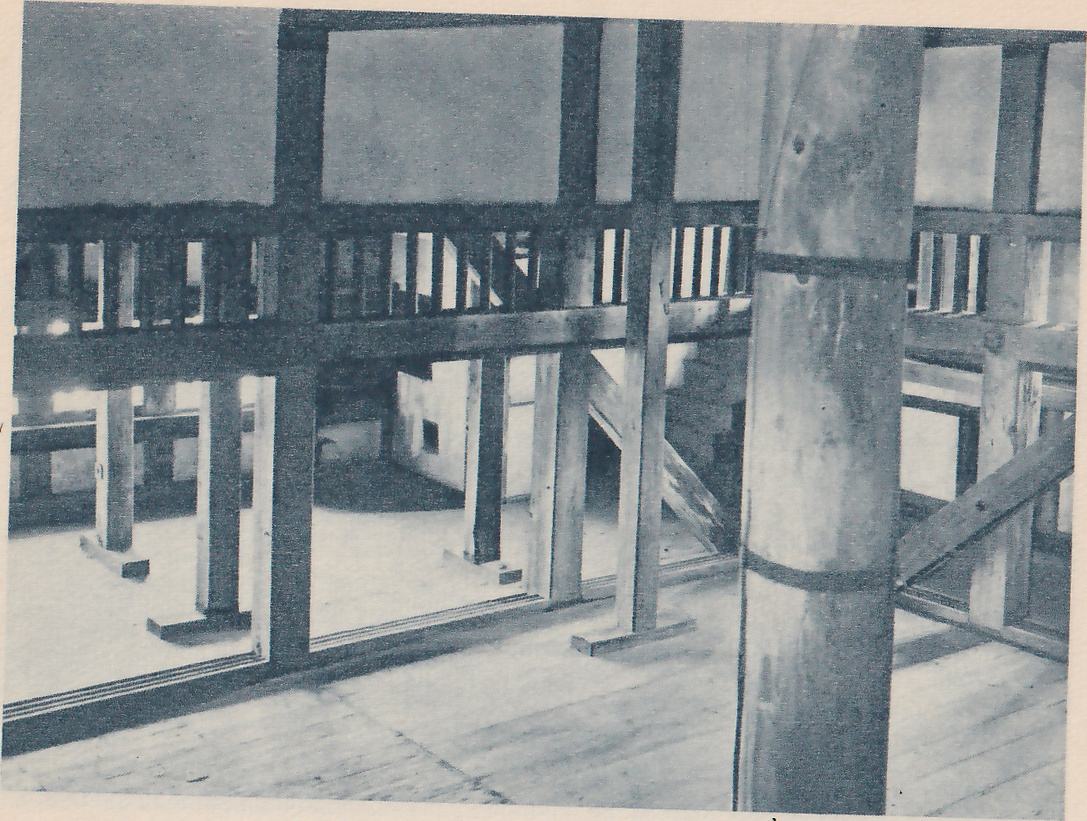 Black-and-white photograph of the inside of a large wooden room, with a thick round post wrapped by two dark bands at the right. Square wooden posts and beams form a frame across the picture, with a row of slats at the upper middle and a floor of boards in the foreground. Pale light comes through openings in the walls, and slanting wooden braces cross at the center and right.