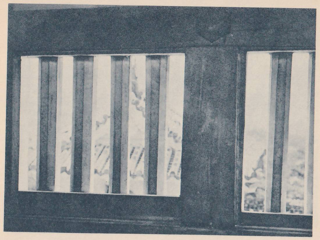 Black-and-white photograph of a dark wooden wall with two window openings side by side, the wider one at the left and a narrower one at the right, each crossed by thick vertical wooden bars with a bright, blurred view between them. A small pointed shape is set into the wall's upper edge above the center.