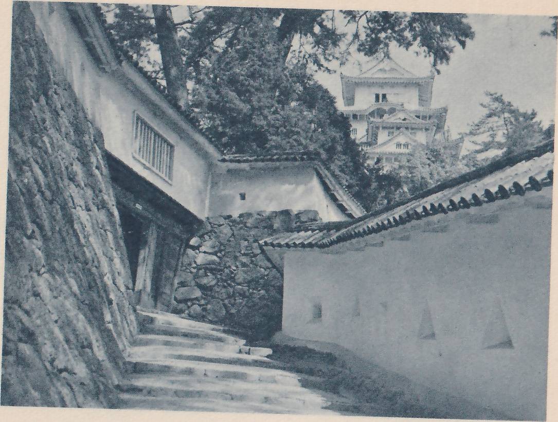 Black-and-white photograph looking up a flight of pale steps that runs between a steep wall of rough stones at the left and a long white wall with a tiled top at the right. A white-walled building with a barred window stands above the steps at the left, with a dark gateway under it. Beyond them a white-walled building of stacked tiled roofs rises above dark trees at the upper right. A few small square and pale triangular openings show in the white wall at the right.
