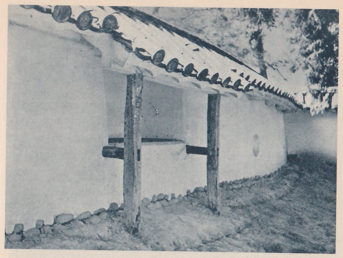 Black-and-white photograph of a curved white wall under a tiled roof edge lined with round tile ends, seen from below at an angle. Two rough wooden posts stand against the wall, with dark horizontal pieces crossing between and beside them. Round stones line the foot of the wall, and a small oval opening shows in the wall at the right. Trees show at the upper right.