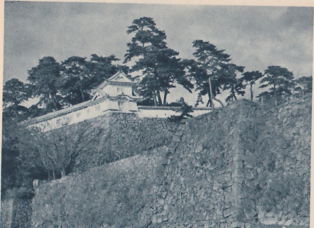 Black-and-white photograph of a steep sloping wall of fitted stones filling the foreground and rising to a long white wall with a tiled top. At the upper left a small white-walled building with a gabled tiled roof stands on the wall. Pine trees of several heights stand along the skyline under a cloudy sky, and a small bush shows at the center.