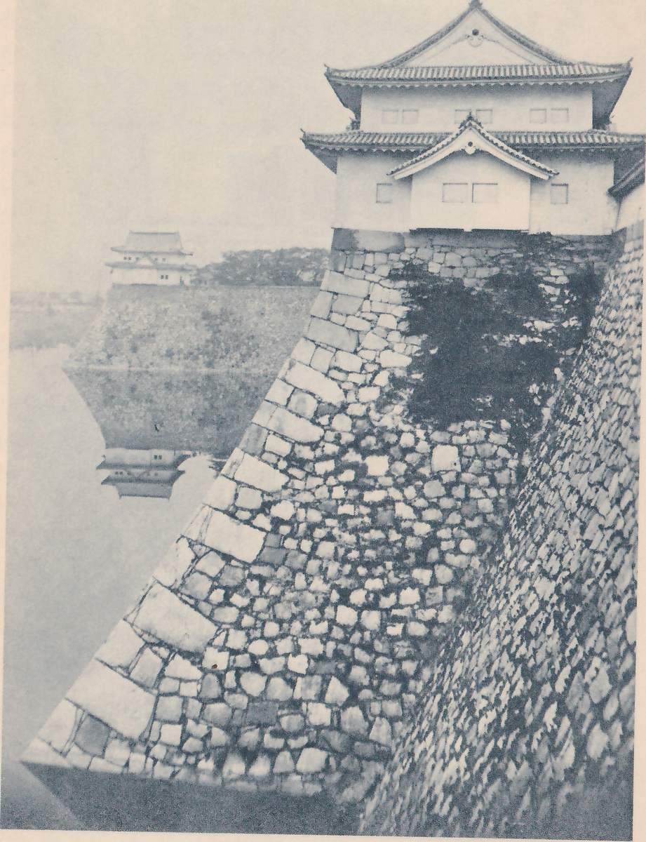 Black-and-white photograph of a high wall of fitted stones that slopes out toward its foot, with a corner of larger squared stones running diagonally from the lower left up toward the center. A white-walled tower with stepped tiled roofs and a gable stands on top at the upper right, and a dark patch of leaves clings to the wall below it. At the left a second wall rises from still water, with a smaller white-walled building on top that is mirrored in the water. A wall of smaller close-set stones runs up the right edge.