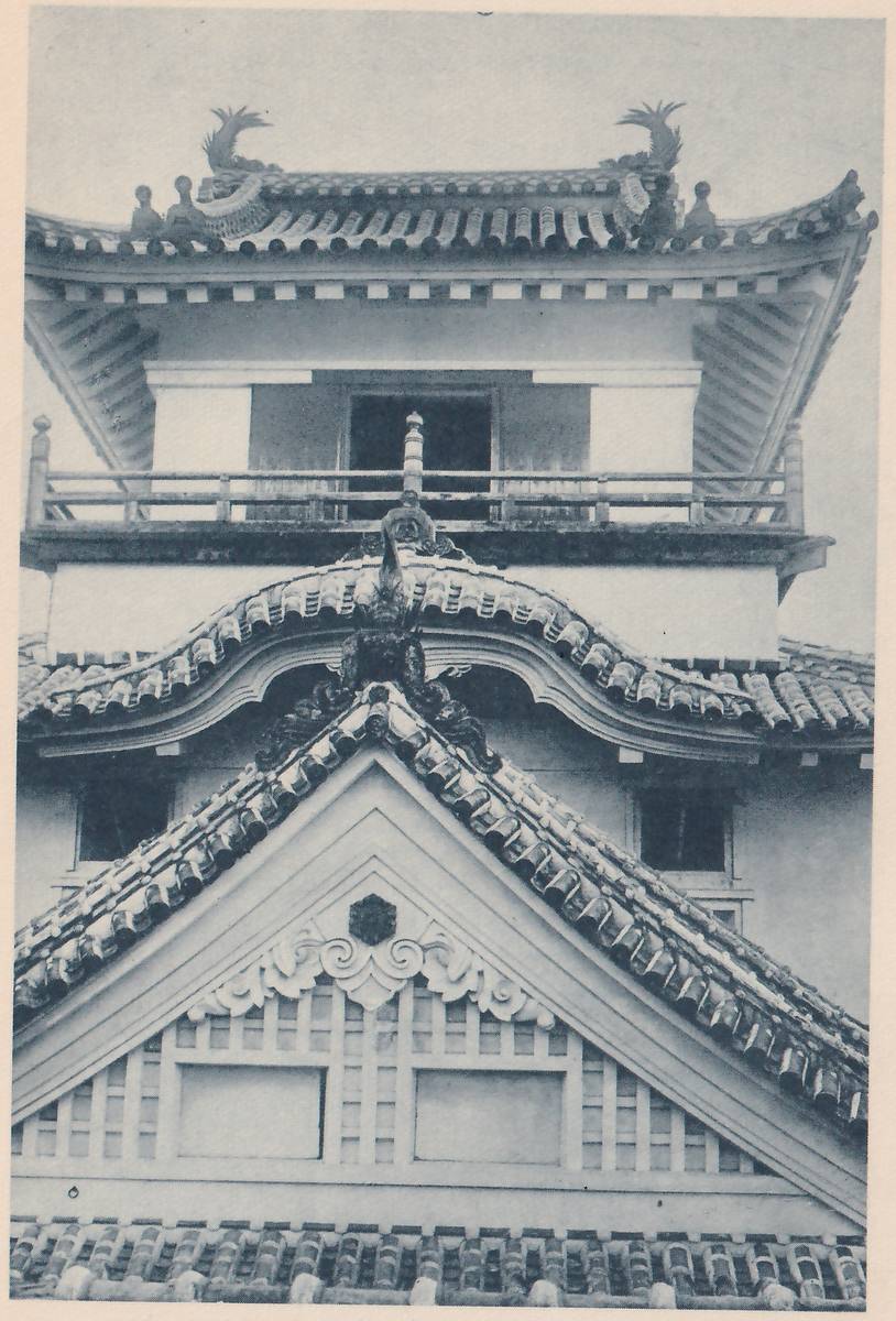 Black-and-white photograph, close up, of the upper part of a building of stacked tiled roofs. A curling, finned ornament stands at each end of the top roof's ridge. Under that roof a railed balcony runs in front of a dark window opening. Below it a curved roof has a dark ornament at the center of its ridge, and below that a large pale triangular gable holds a lattice panel, scrolled carving and a small hexagon-shaped mark at its peak. Rows of tiles with round ends fill the bottom edge.
