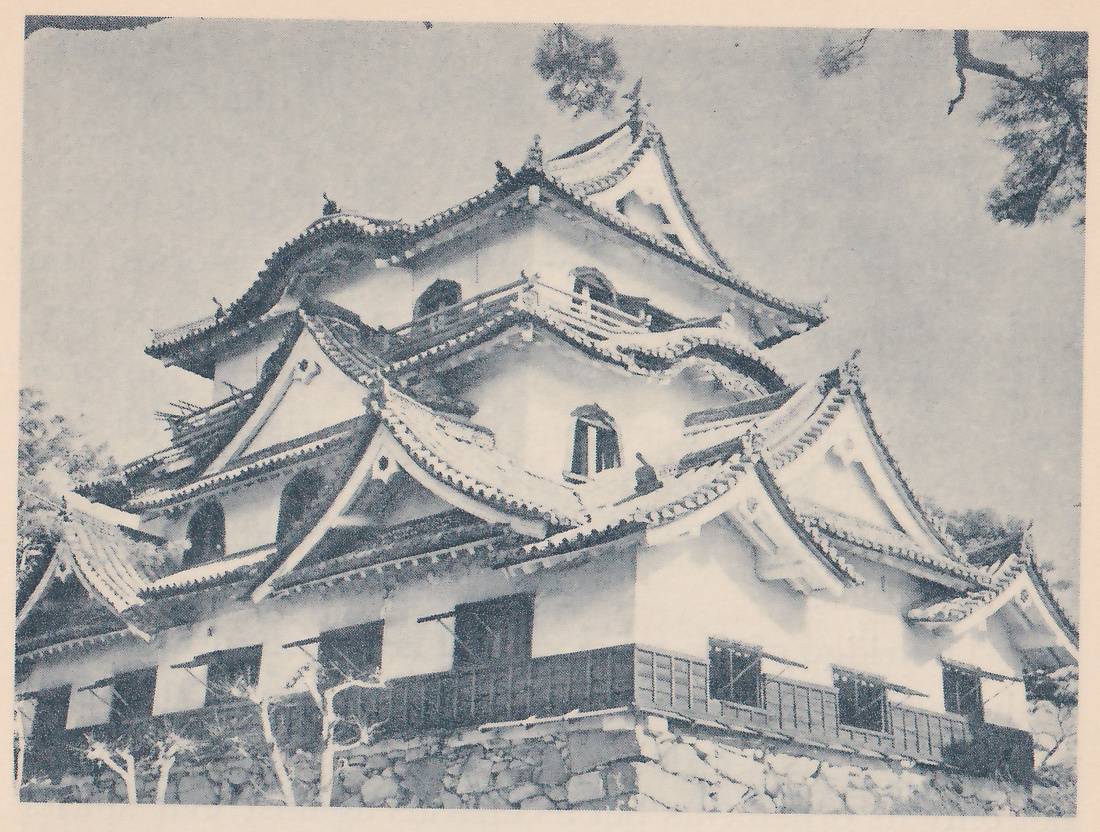 Black-and-white photograph, taken from below, of a white-walled building of stacked roofs, with many curved gables and wavy-edged roofs layered around its upper part. Arched openings and small windows show in the upper walls, a short railing runs along a middle level, and a dark band of wooden panels with square windows and small slanting awnings runs along the base above a wall of fitted stones. Pine branches show at the upper corners.
