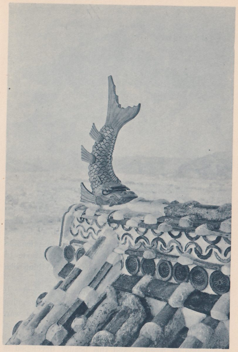 Black-and-white photograph, close up, of a roof corner topped by a fish-shaped ornament with a scaled body, fins and an open-mouthed head, its tail curving high up and back. Below it a band with a wave pattern and round tile ends runs along the roof edge, and rows of tiles run down a slope at the lower left. A pale hazy ground and distant hills lie behind.