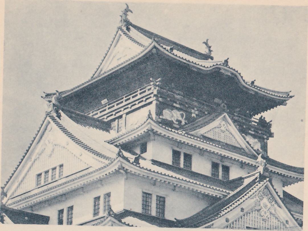 Black-and-white photograph, taken from below, of the upper part of a building of large stacked roofs against the sky. Curved gables with fine lattice panels and rows of small windows stand at the left and lower right, pointed ornaments stand at the ridge ends, and a railing runs along a middle level. On the dark wall below the top roof are pale figures, one near the center and one at the right.