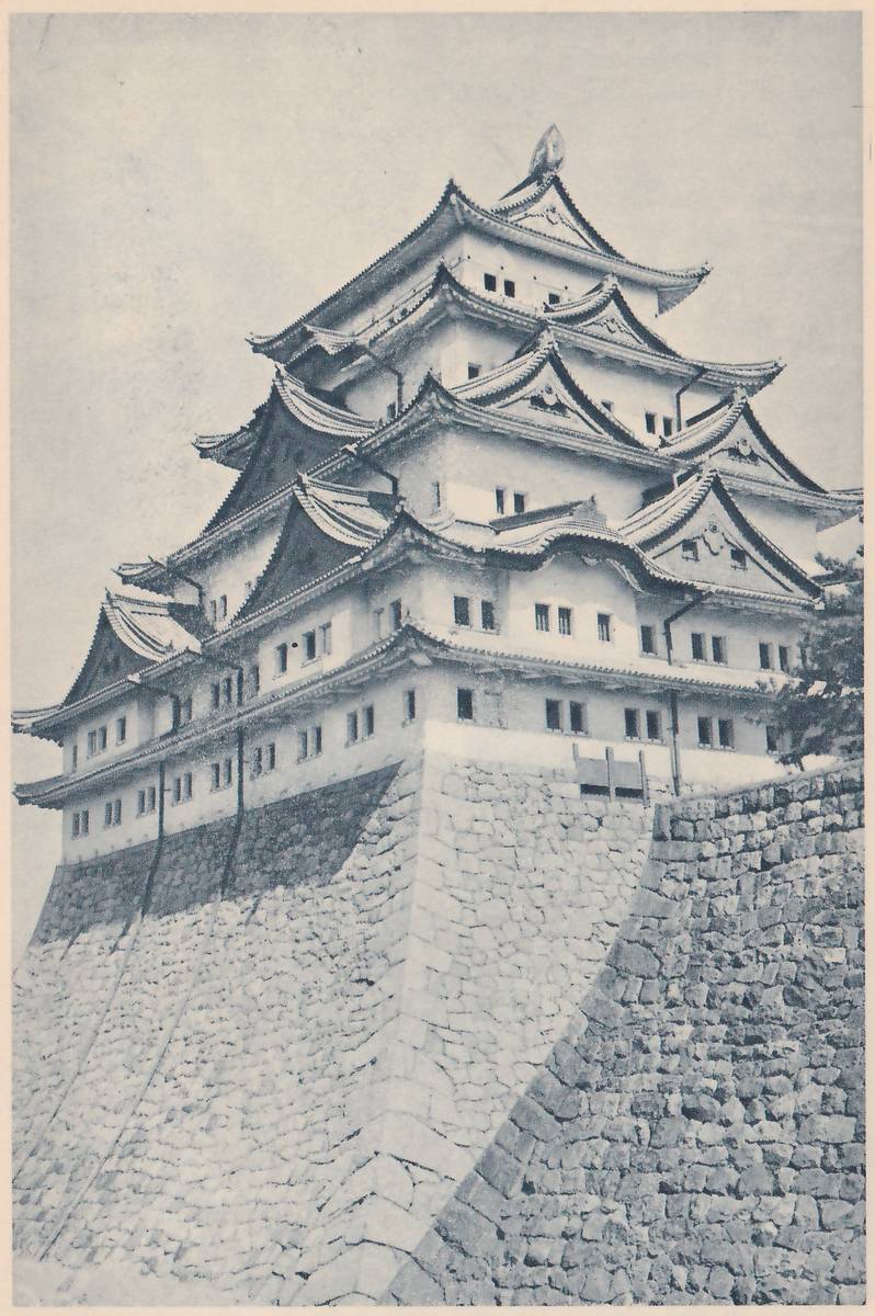 Black-and-white photograph, taken from below, of a tall white-walled building of stacked tiled roofs and gables with rows of small windows and an ornament at its very top, standing on a high base whose stone wall slopes steeply toward the lower left. A second wall of close-set stones rises in front at the lower right, and a tree shows at the right edge.