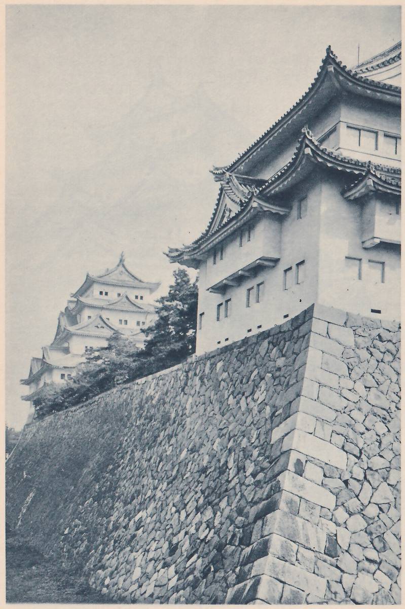 Black-and-white photograph of a white-walled building with several stacked roofs, gables and rows of small windows at the right, standing on a tall sloping wall of fitted stones whose squared corner stones rise in a ridge. At the left, farther off, a taller building of stacked roofs shows above trees against a hazy sky.
