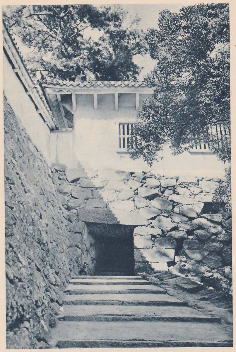 Black-and-white photograph looking up a flight of broad flat steps toward a dark opening cut through a wall of large fitted stones, with a steep rough stone wall rising at the left. Above the opening a white-walled building with a tiled eave and barred windows stands on the stones, and leafy branches hang over from the right and top. A white wall with a tiled top runs along the upper left.