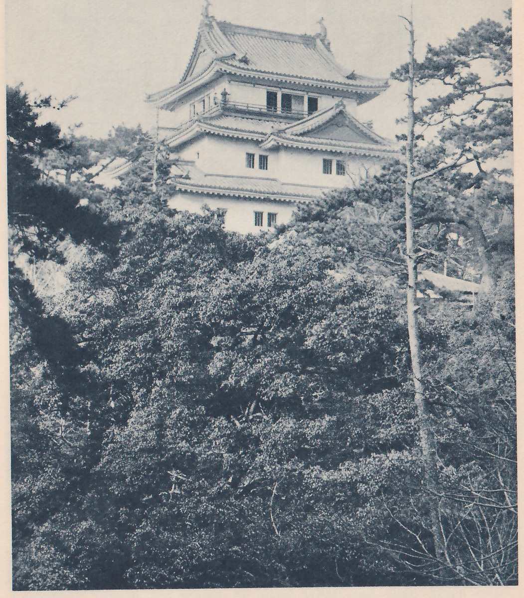 Black-and-white photograph of a white-walled building with stepped tiled roofs and a railed upper level with dark openings, rising above dense leafy trees that fill the lower three-fifths of the picture. A bare slender pine trunk stands at the right, and the corner of a low roof shows among the trees at the right edge.