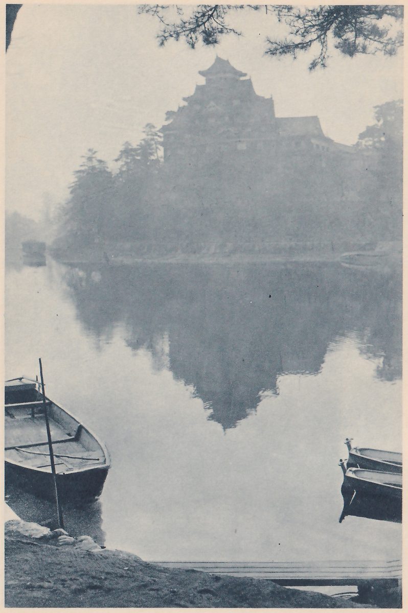 Black-and-white photograph in mist of still water, with a large dark building of stacked roofs rising behind a bank of trees on the far side and its dim reflection spreading down the water toward the viewer. A boat with a pole standing upright in it lies at the lower left, two more boats lie close together at the lower right, and a dark bank and a flat plank edge show at the bottom. Pine branches hang at the upper right.
