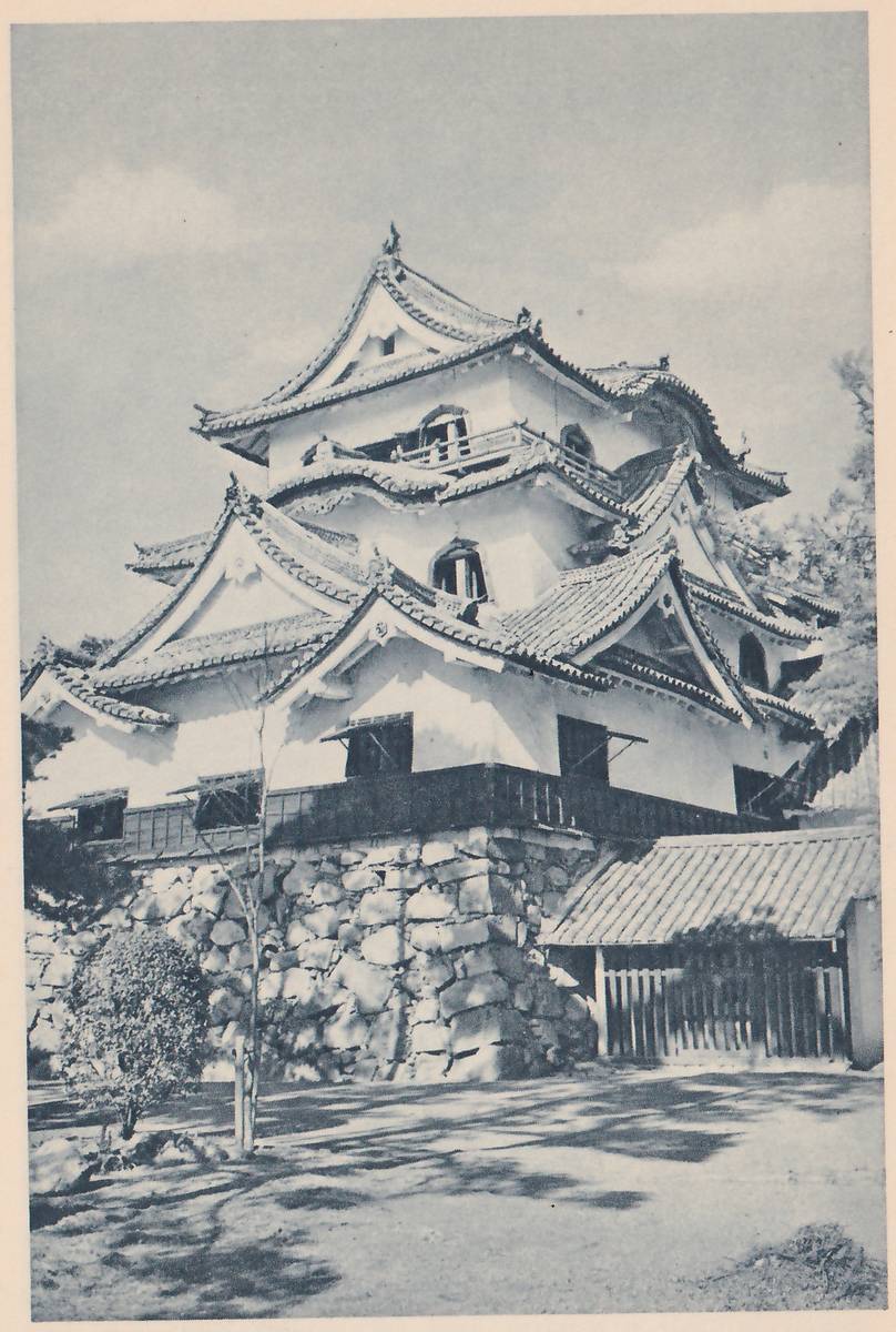 Black-and-white photograph, taken from below, of a white-walled building of stacked roofs with curved gables, arched windows and a short railing on an upper level, standing on a base of fitted stones with a dark band of wooden panels and awning-like window hoods around it. At the right a low tiled-roof gateway with a barred wooden door stands beside the stones. Shadows of branches cross the open ground in front, and a young tree and a clipped shrub stand at the left.