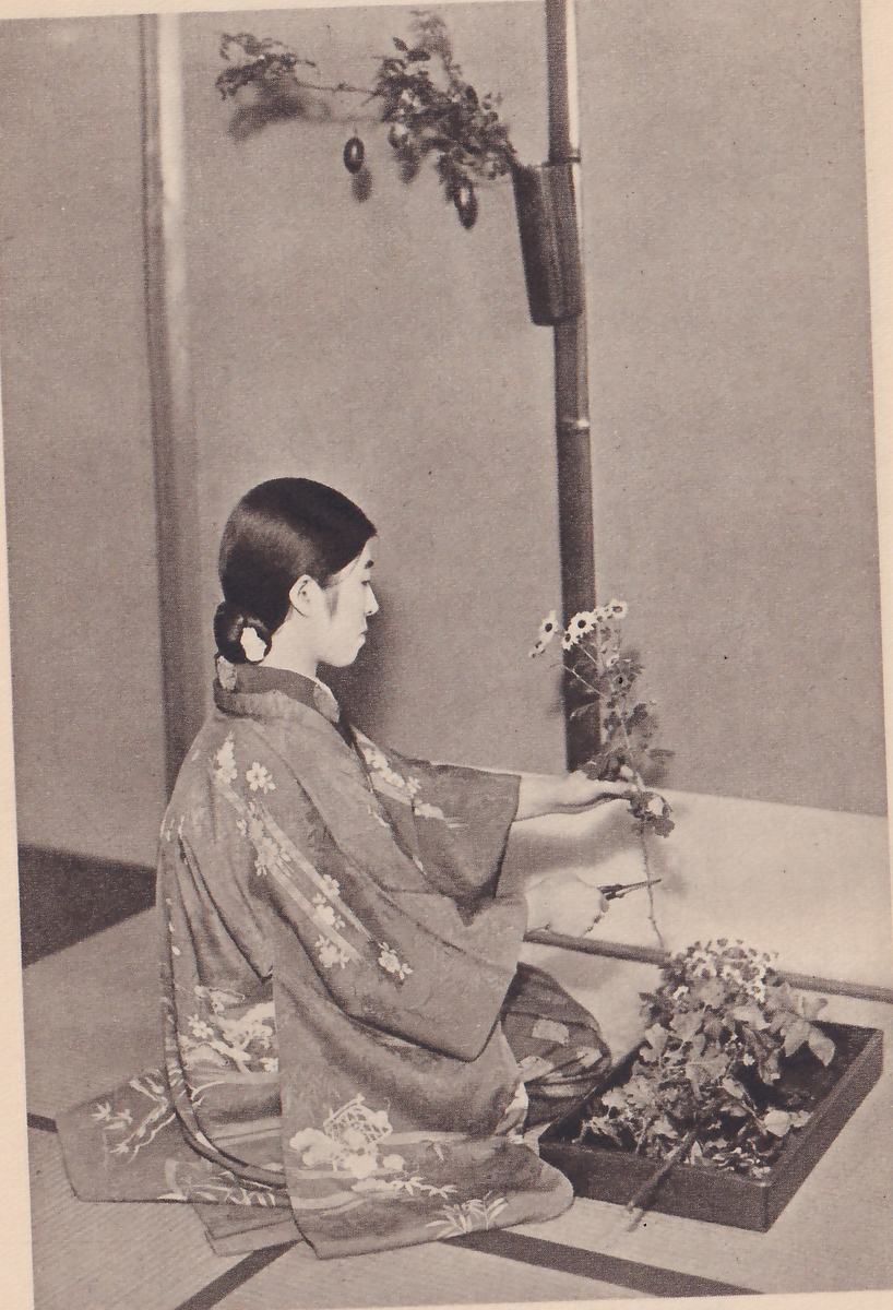 Photograph of a young woman in a flowered kimono kneeling in profile facing right, holding a stem of small white-petaled flowers and a pair of small scissors; a hanging vase with an arrangement is above her, and a dark flower tray lies on the floor at the right.