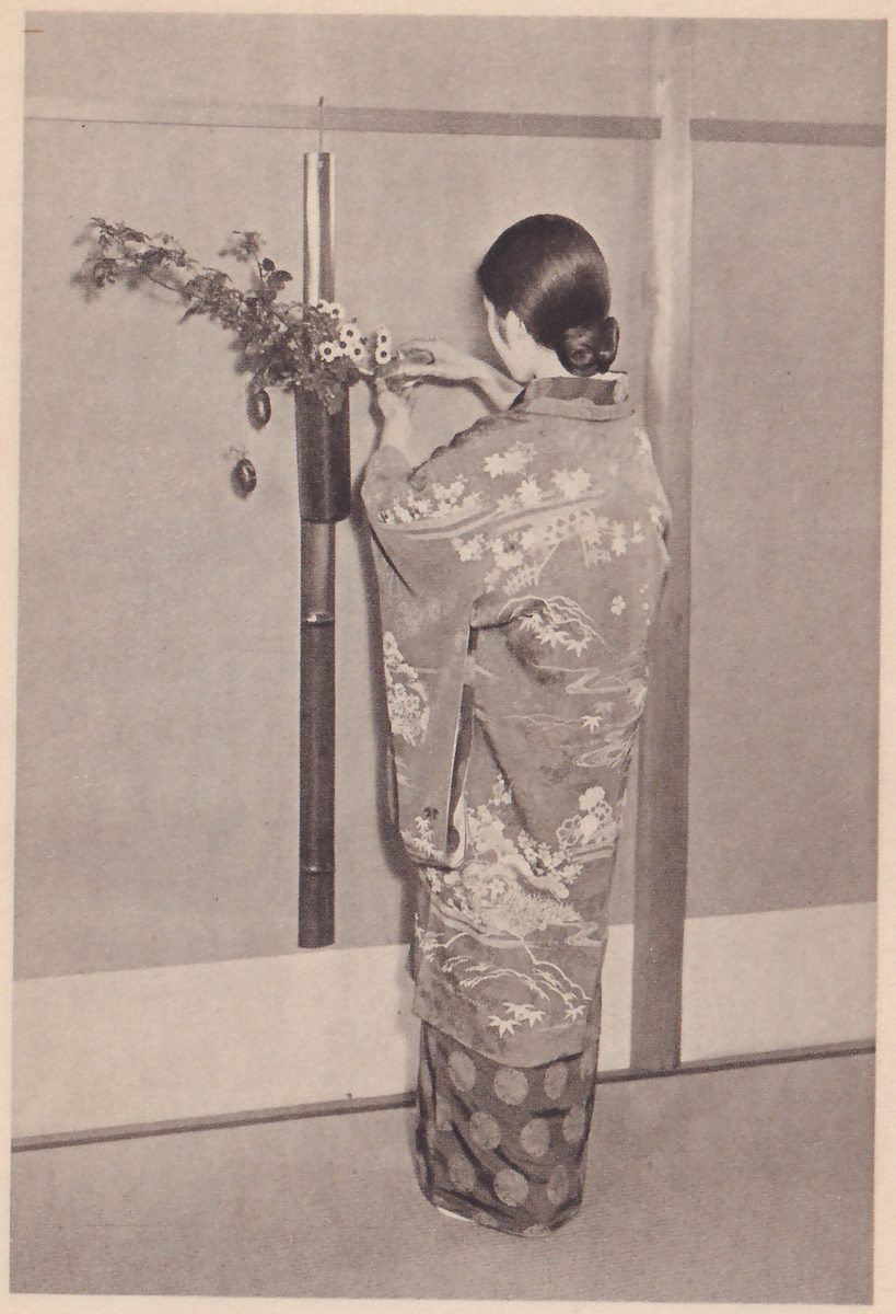Photograph of the woman in the maple-leaf kimono, seen from behind, with her hands raised to a hanging bamboo vase that holds a branch with leaves, small white flowers and a small hanging oval fruit.