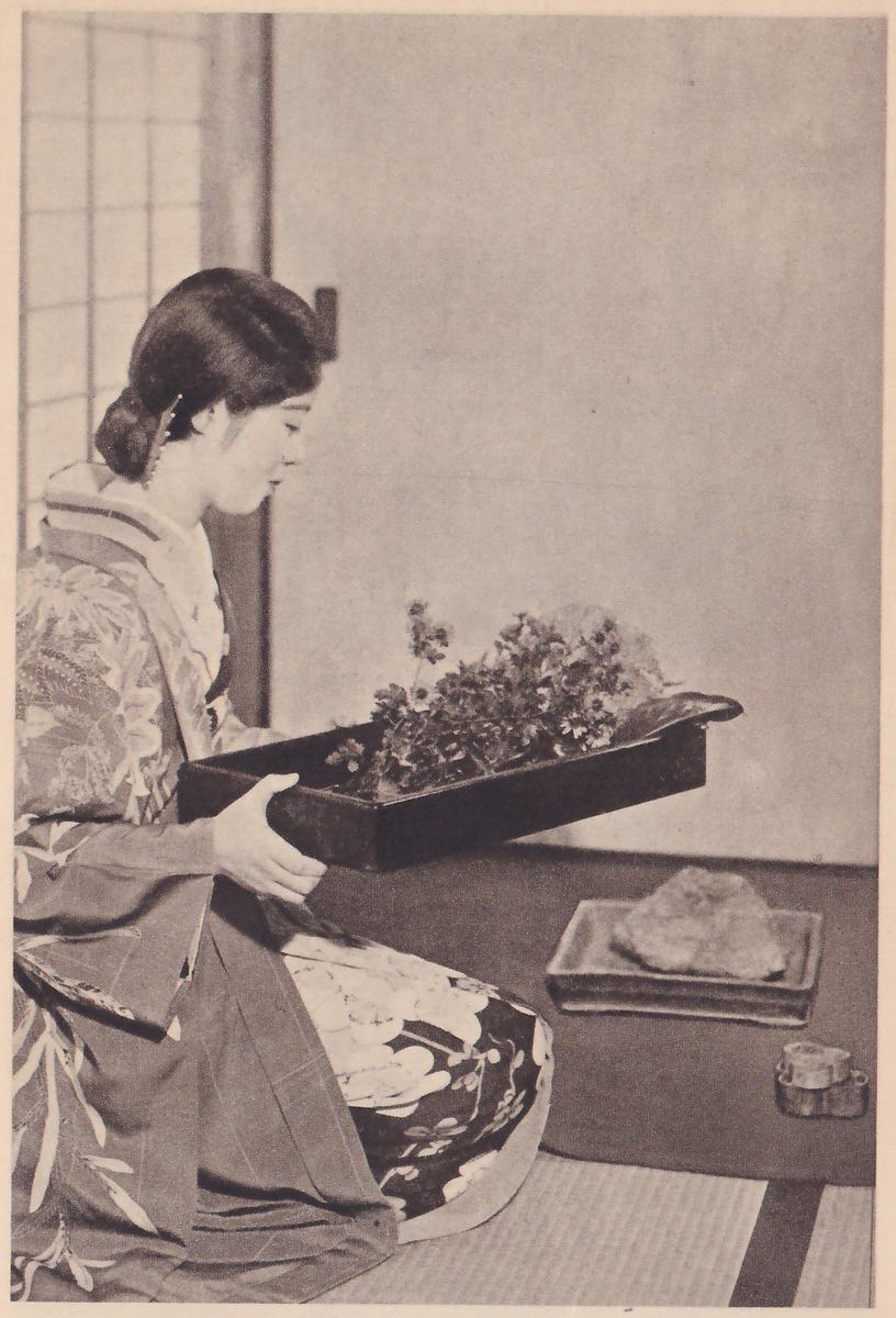 Photograph of a young woman in a flowered kimono kneeling in profile facing right and carrying a dark flower tray of small blossoms and leaves in both hands; on the floor in front of her are a shallow dish with a rock in it and a small flower-holder.