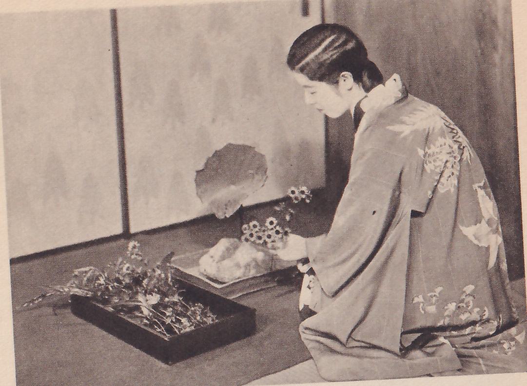 The upper of two photographs: a young woman in a kimono with a pattern of leaves, kneeling in profile facing left, sets a flower beside a rock in a shallow dish, a large round leaf rising behind the rock; a dark tray of flowers lies on the floor in front of her.
