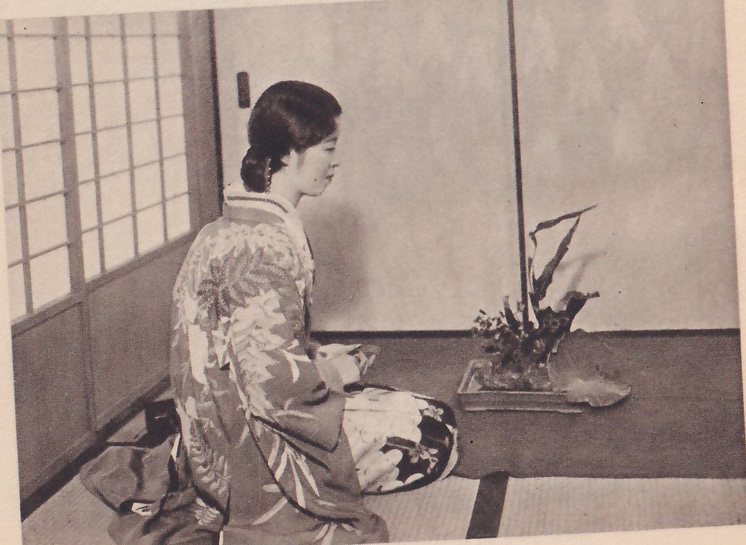 The lower of two photographs: a young woman in a kimono with a pattern of large leaves, kneeling in profile facing right in front of a window of paper panes, with the finished arrangement of leaves and flowers in the shallow dish on the mat beside her.