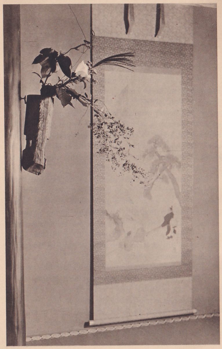 Photograph of a tokonoma wall: a hanging scroll with a painting of a tree, rocks and a small figure, and beside it, on the pillar at the left, a vase of dark wood holding leaves, a white flower and long sprays of small blossoms that trail across the scroll.