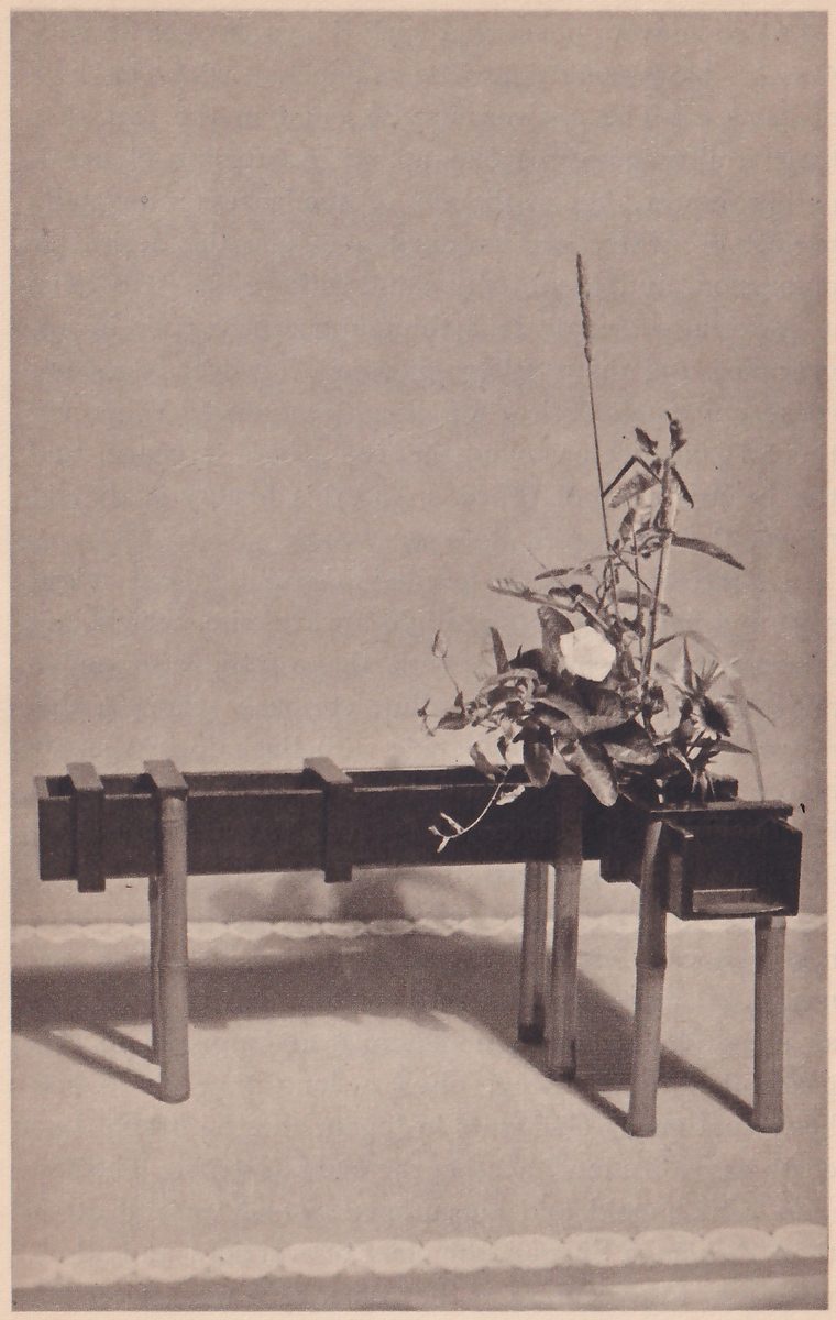 Photograph of a long narrow dark wooden trough-shaped vase on four bamboo legs, with a spout box at its right end; at the right end rise a white flower, leaves and a tall slim grass spike, and the left end of the trough is bare.