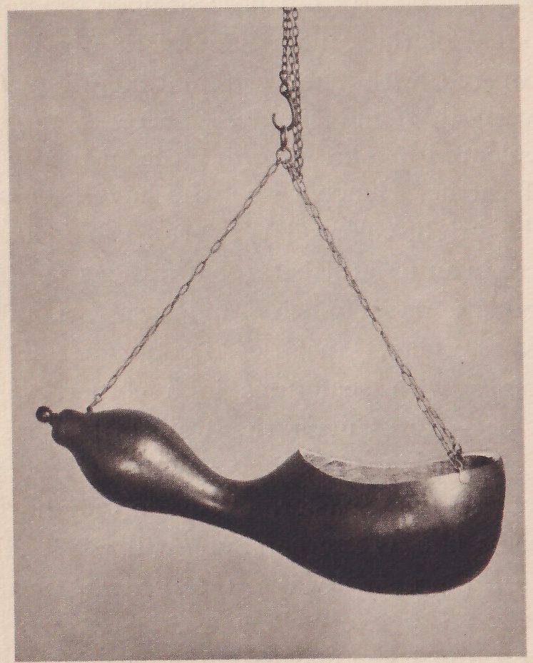 Photograph of a dark gourd hung by two chains from one hook, lying nearly level, its narrow neck at the left and a wide opening cut in the top of its bulb at the right, against a plain gray ground.