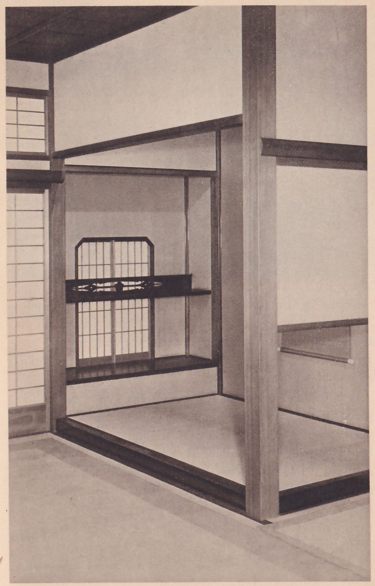 Photograph of a plain Japanese room seen from a corner: at the center a raised alcove with a low floor, a window with sliding paper panes and a shelf with a carved front, a dark wooden post at the right.