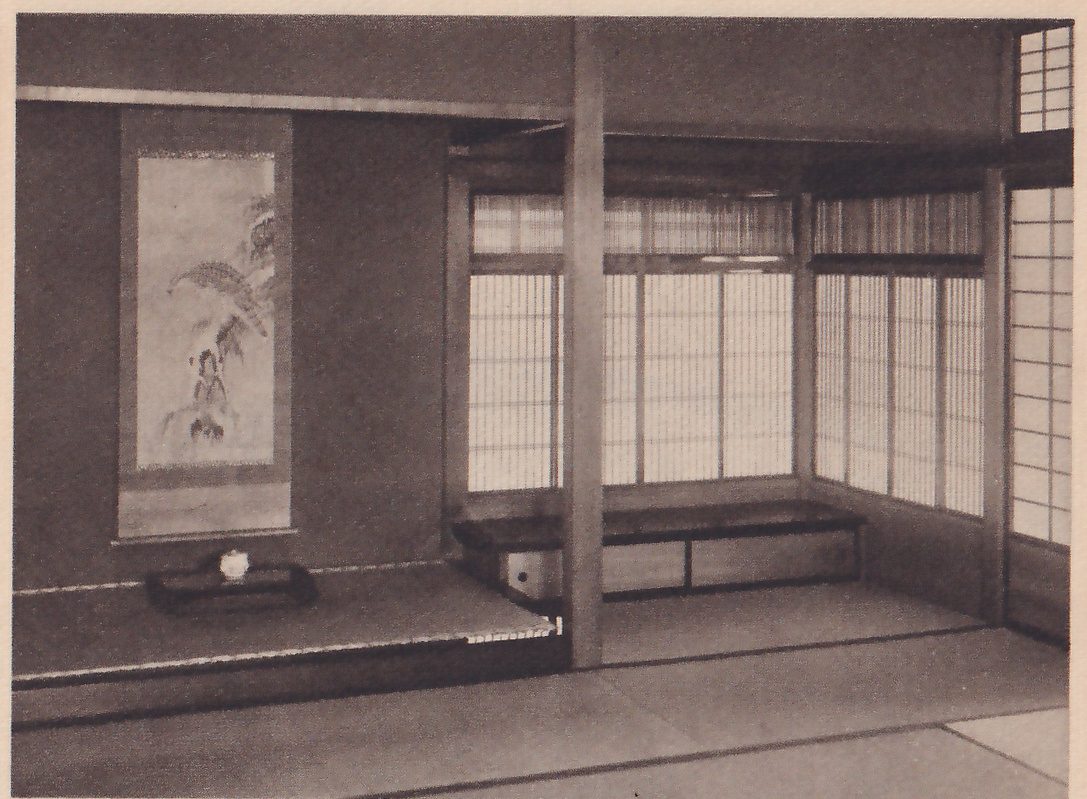 Photograph of a tokonoma in a mat room: at the left a hanging scroll painted with a leafy plant above a low tray holding a small white pot, and at the right a row of sliding paper windows with a long low desk-like shelf below them.