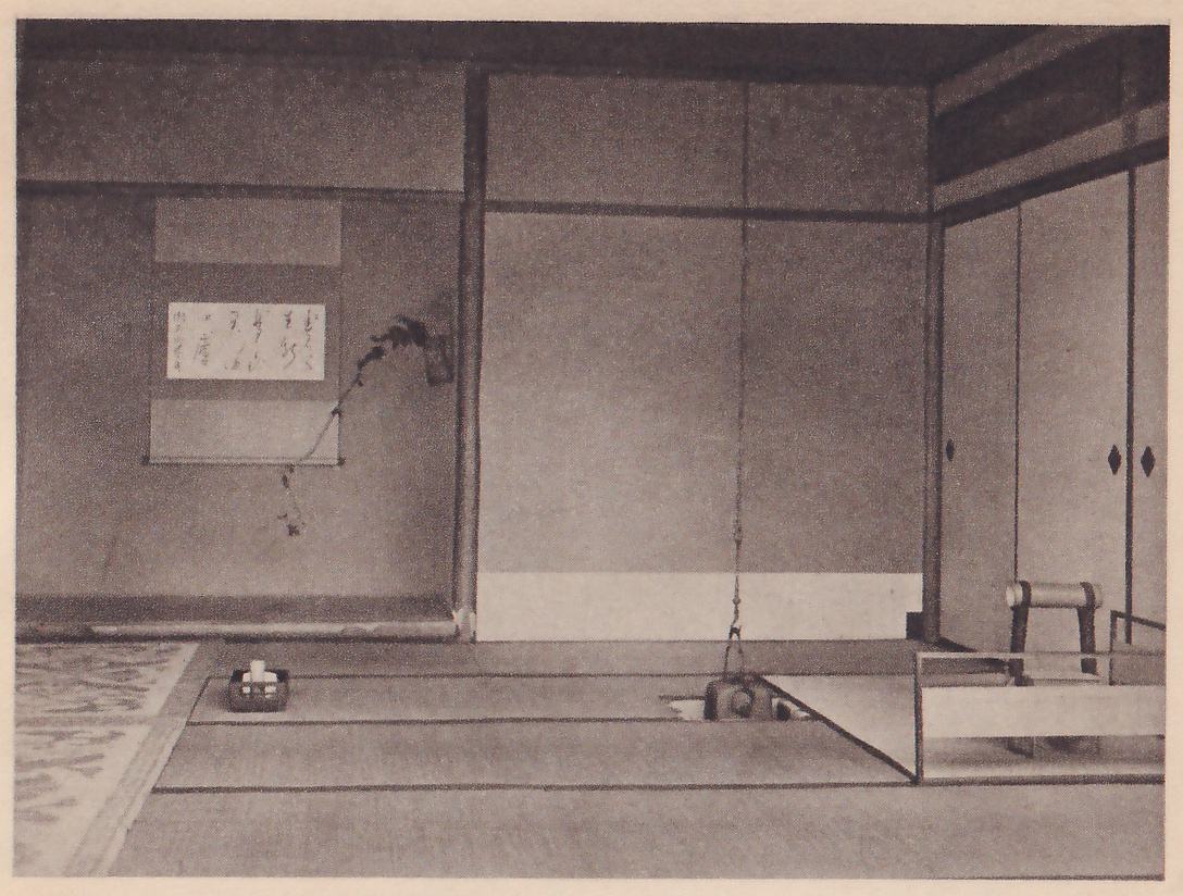 Photograph of a mat room with, on the far wall, a hanging scroll of a few lines of writing and a hanging vase on the post beside it with a long spray of leaves trailing down; on the floor a small basket and a sunken hearth with a chain hanging above it, and a low screen at the right.