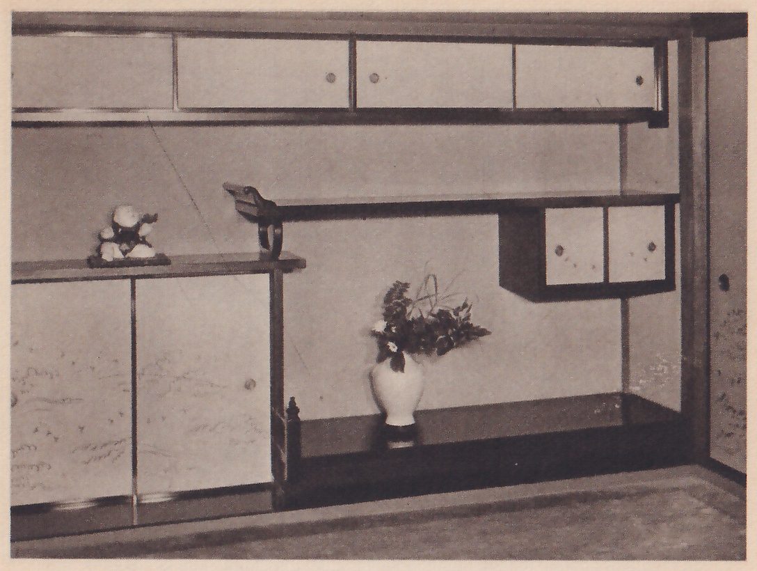 Photograph of a wall of shelves in a room: a long upper cabinet with three sliding doors, a long shelf under it, a lower cabinet with painted doors at the left holding a small figure on top, and a lower floor level at the right with a white vase of flowers on a stand.