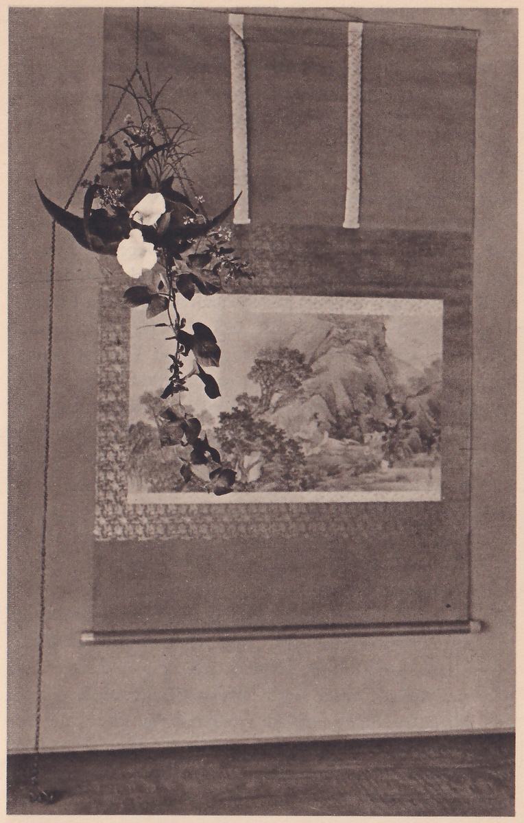 Photograph of a large hanging scroll of a mountain landscape with a vase of white flowers and leaves hung in front of it by a thin string, the flowers falling in a line from the top across the picture.