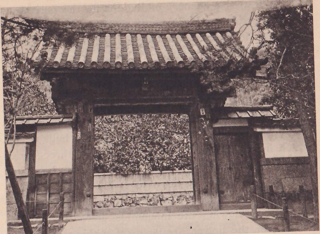 Photograph of a wooden temple gate with a tiled roof seen from the front, an open doorway at the center showing a bamboo fence and bushes beyond, a plain white panel and a closed door on either side, and trees at the edges.